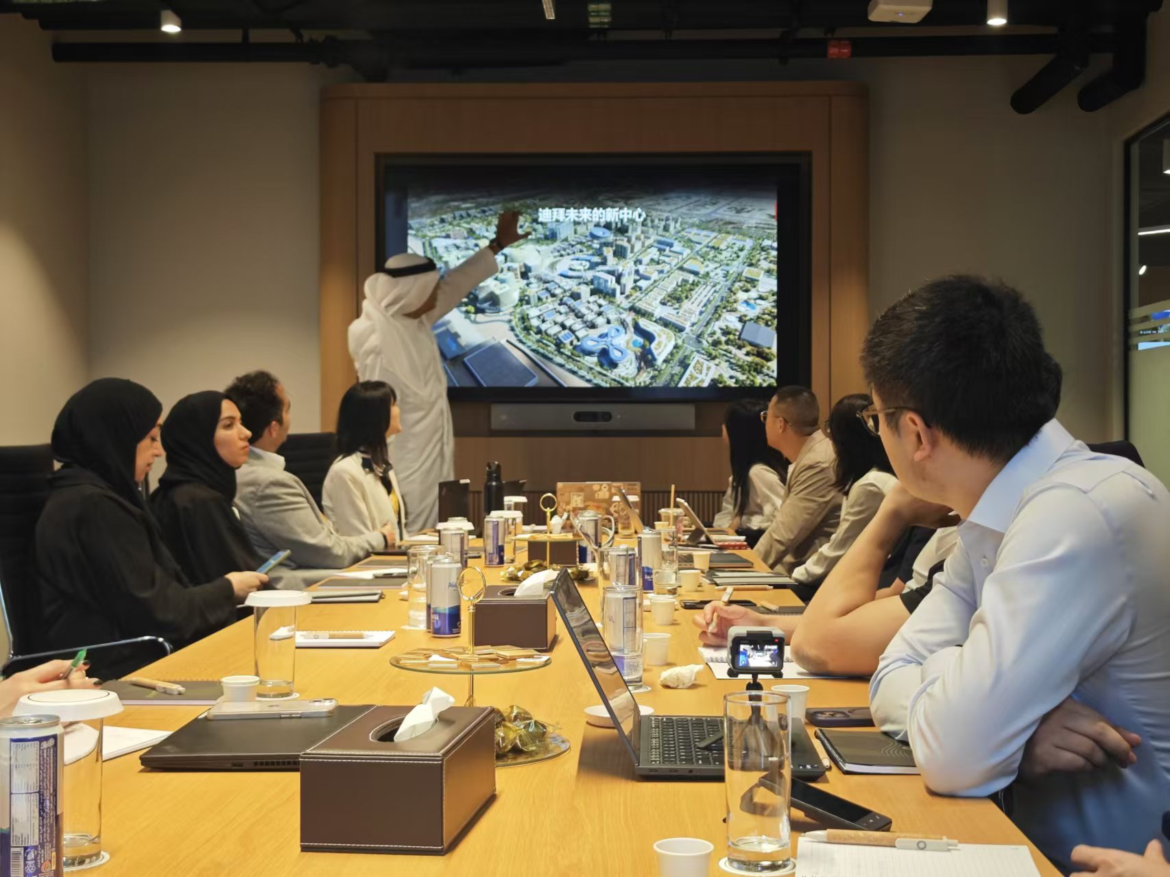 In a warmly lit conference room, a man in traditional emirati attire gestures passionately toward a large screen displaying the visionary blueprint of 'dubai’s new centre. ' around the table, diverse professionals lean in with focused eyes—some taking notes, others sipping coffee—united by shared purpose and the quiet excitement of shaping tomorrow’s skyline together